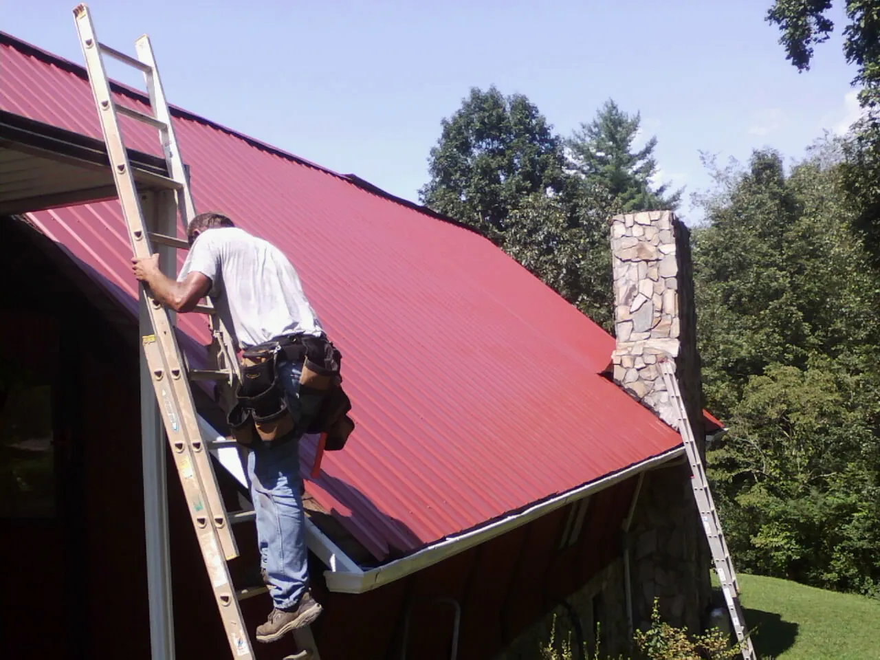 Roofer on a ladder beside a red metal roof and stone chimney — Soffit & Fascia Repair by Roofing Brothers in Somis